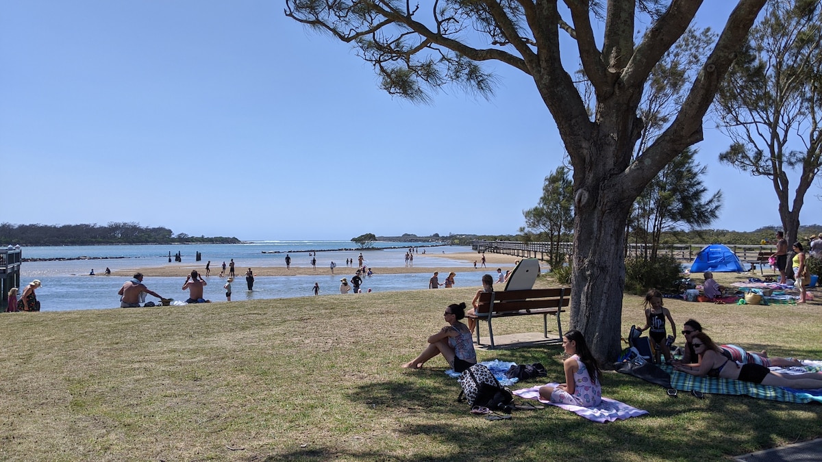 A sandy beach area is visible, where a variety of people enjoy the water. Toward the background, a calm river meets the sea, with children playing. A grassy area is occupied by several guests relaxing on blankets under the shade of trees, creating a peaceful atmosphere.