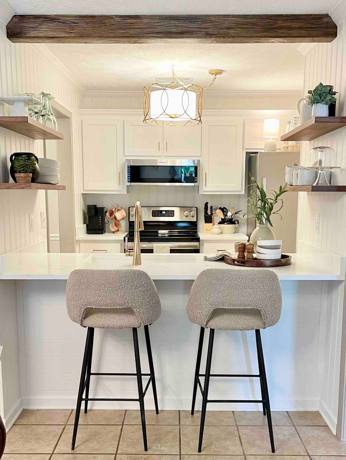 A contemporary kitchen space features a bar area with two upholstered stools. Open shelves hold decorative items and plants, while modern light fixtures hang above. Stainless steel appliances are highlighted in the background, and a quartz countertop offers a clean and bright aesthetic.