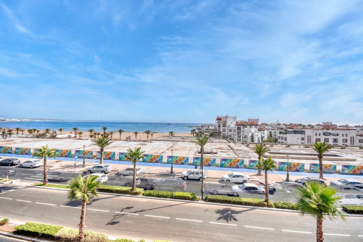A stunning view of the coastline is captured, featuring palm trees lining the avenue. The sea is visible in the background, and the beachfront is framed by colorful artwork along a barrier. Vehicles are parked along the road, contributing to the lively atmosphere.