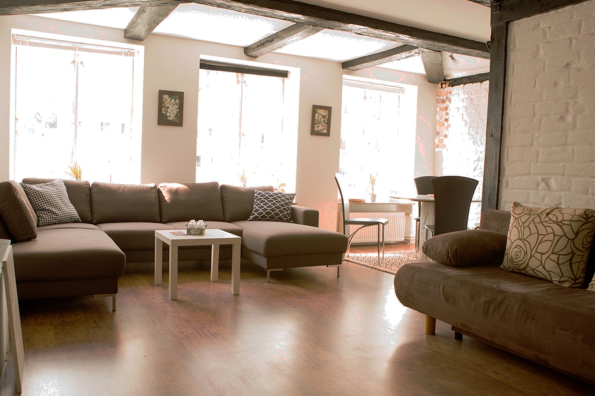 The spacious living area features large windows allowing natural light to fill the room. A comfortable sectional sofa is complemented by a smaller couch with decorative cushions. A white coffee table sits centrally, while a dining table and chairs are visible in the background.
