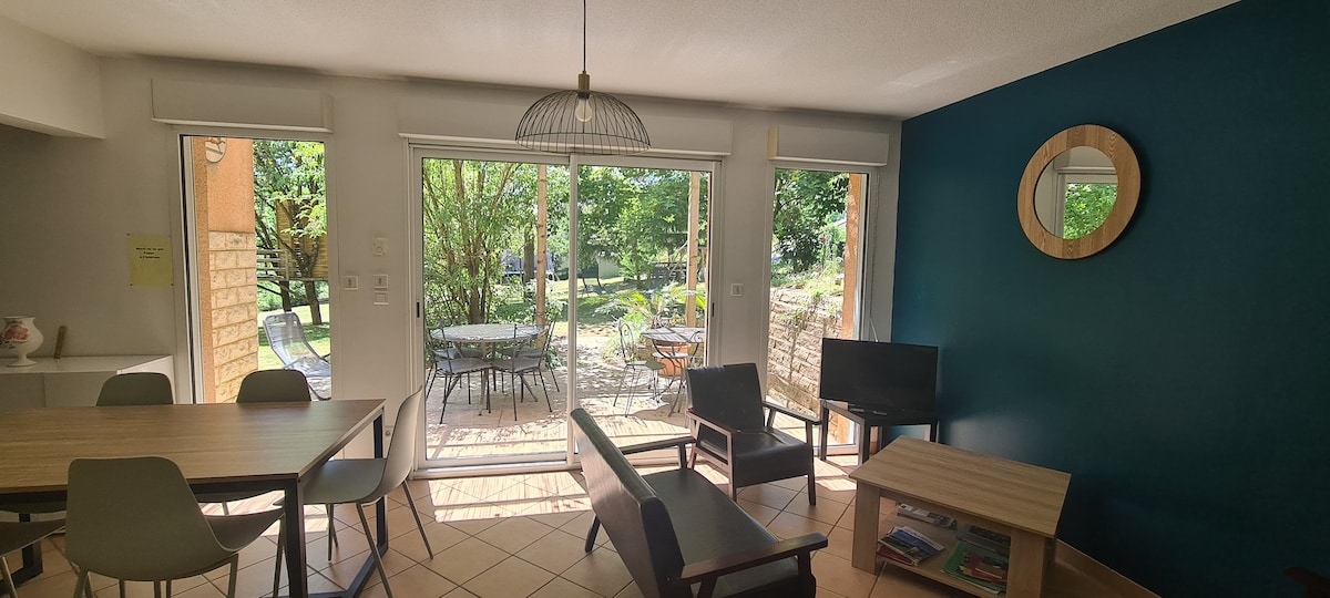 The living area features a wooden table surrounded by chairs, with a comfortable seating arrangement including a sofa and armchair. Large glass doors open to the garden, where outdoor furniture is visible. A round mirror and a light fixture add subtle details to the space.