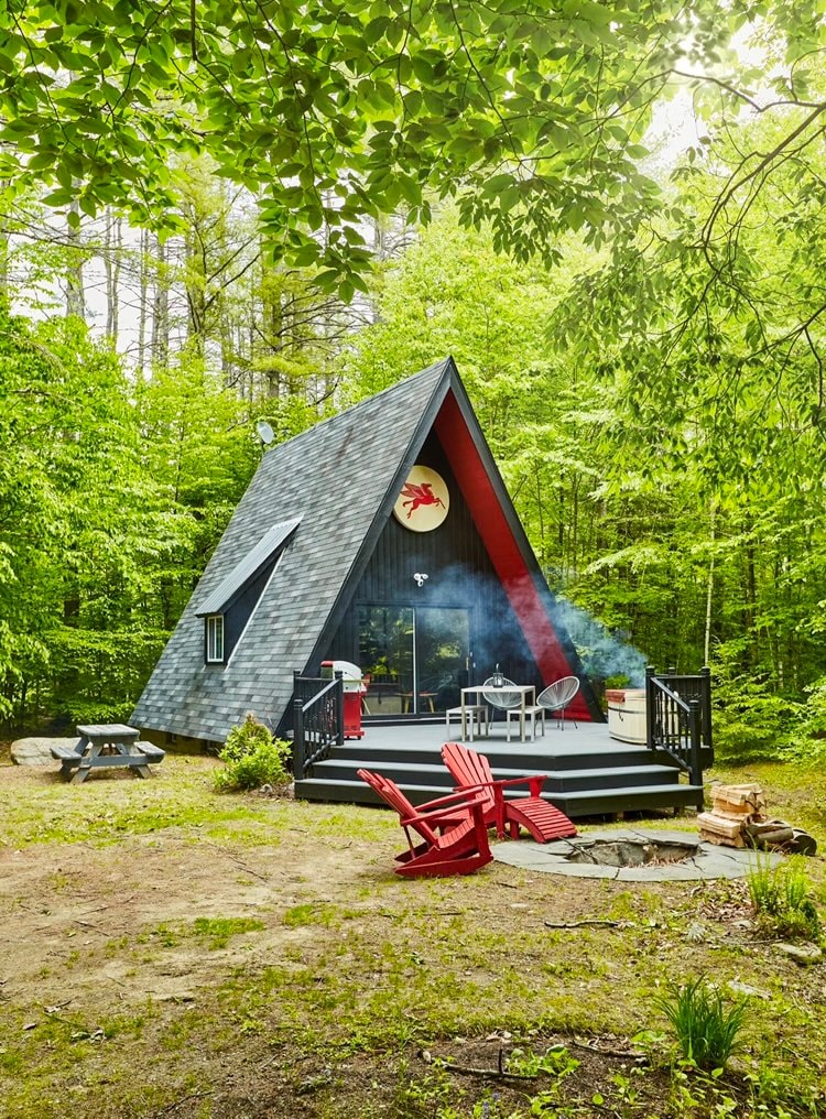 The A-frame cabin is surrounded by lush greenery, featuring a dark exterior with a distinctive red accent. A welcoming deck displays outdoor seating and a fire pit area, while smoke curls gently from the chimney, adding to the tranquil atmosphere.