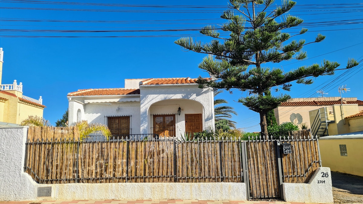 A white bungalow is surrounded by a natural wood fence, complemented by a large pine tree. The exterior is brightened by the clear blue sky above, with a paved walkway leading to the entrance, framed by lush greenery.
