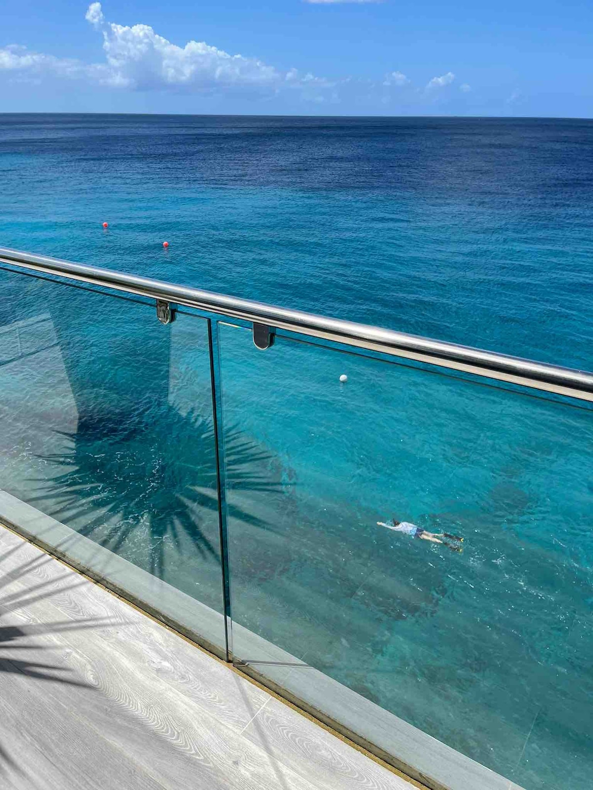 A clear view of the ocean is presented beyond a sleek glass railing, with vibrant blue waters visible below. The sun casts reflections on the surface, while a snorkeler is seen exploring the underwater scenery. A hint of greenery is observed in the foreground.