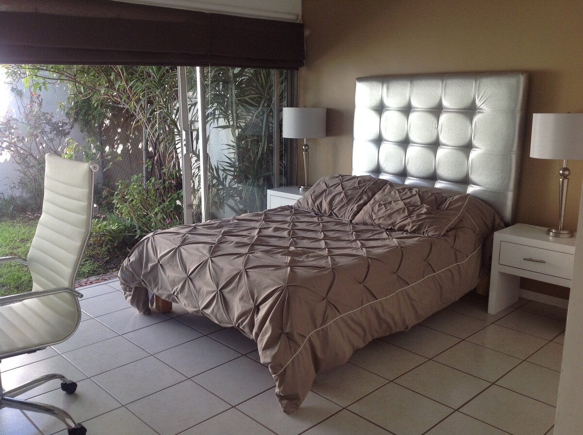 The room features a spacious bed with a tufted silver headboard, dressed in a beige coverlet. Two bedside tables with lamps flank the bed, while a modern white chair is positioned nearby. Natural light enters through sliding glass doors that lead to a green garden space.