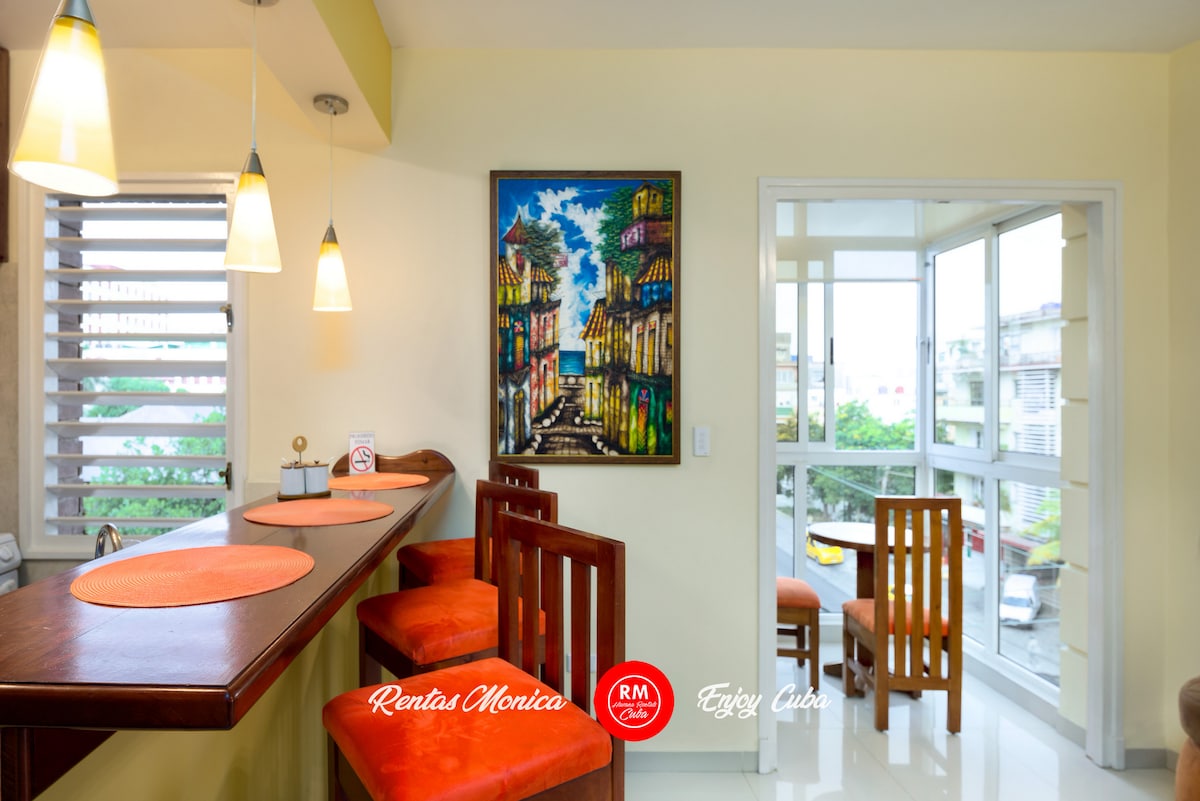A well-lit dining area features a wooden bar with orange placemats and four wooden chairs. A vibrant painting depicting a Cuban street scene adorns the wall. Large windows provide a view of the balcony and natural light floods the space.