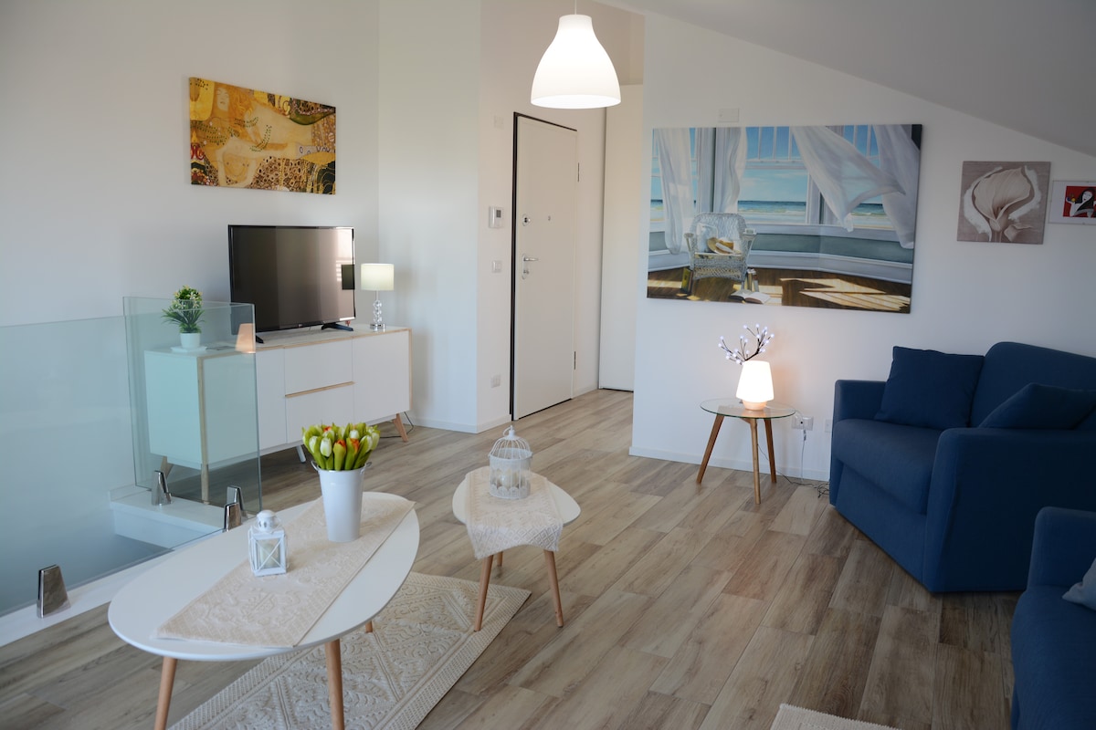 A contemporary living area is featured with light wooden flooring and large windows allowing natural light. A blue sofa sits beside a stylish coffee table adorned with decorative items. A wall-mounted television and artworks add visual interest to the space.