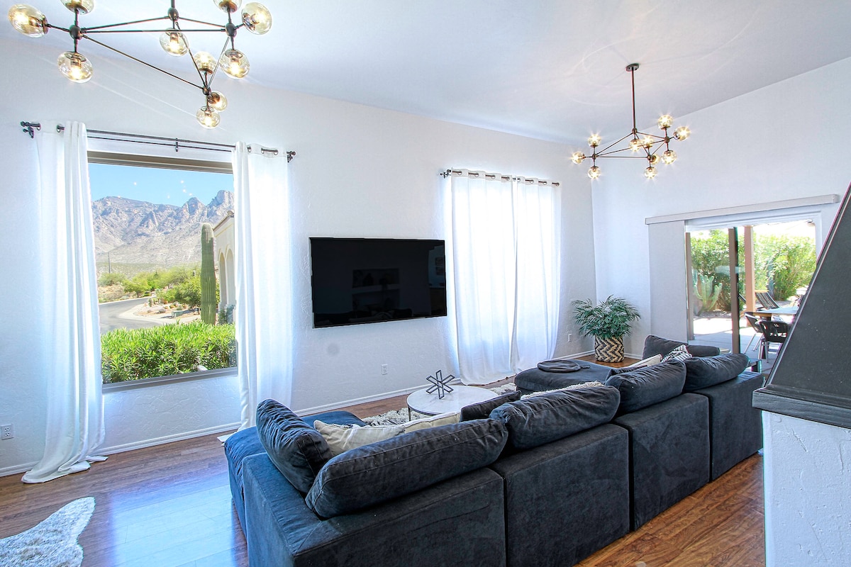 A spacious living area is highlighted by large windows framing stunning mountain views. A comfortable sectional sofa and a modern television are visible, with natural light filtering through sheer curtains. Unique chandeliers provide illumination, complementing the bright and open atmosphere.