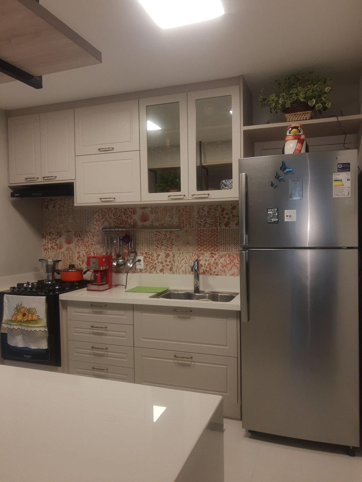 A bright kitchen area is equipped with modern appliances, including a sleek stainless steel refrigerator and a stove. The cabinetry features a light finish, complemented by an eye-catching patterned backsplash. Counter space is available for meal preparation, with utensils and appliances neatly arranged.