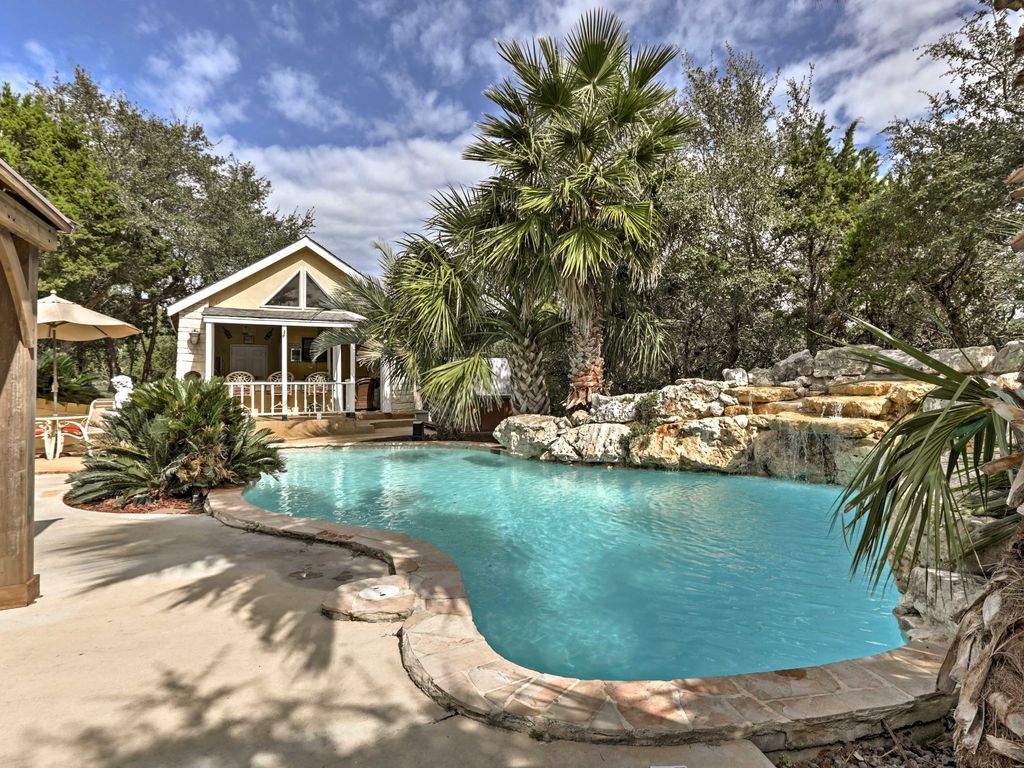A natural lagoon-style pool is surrounded by tropical landscaping and rock formations. A small waterfall flows into the pool, with a spacious deck area that includes lounge chairs and an umbrella. A charming house is visible in the background, framed by trees.