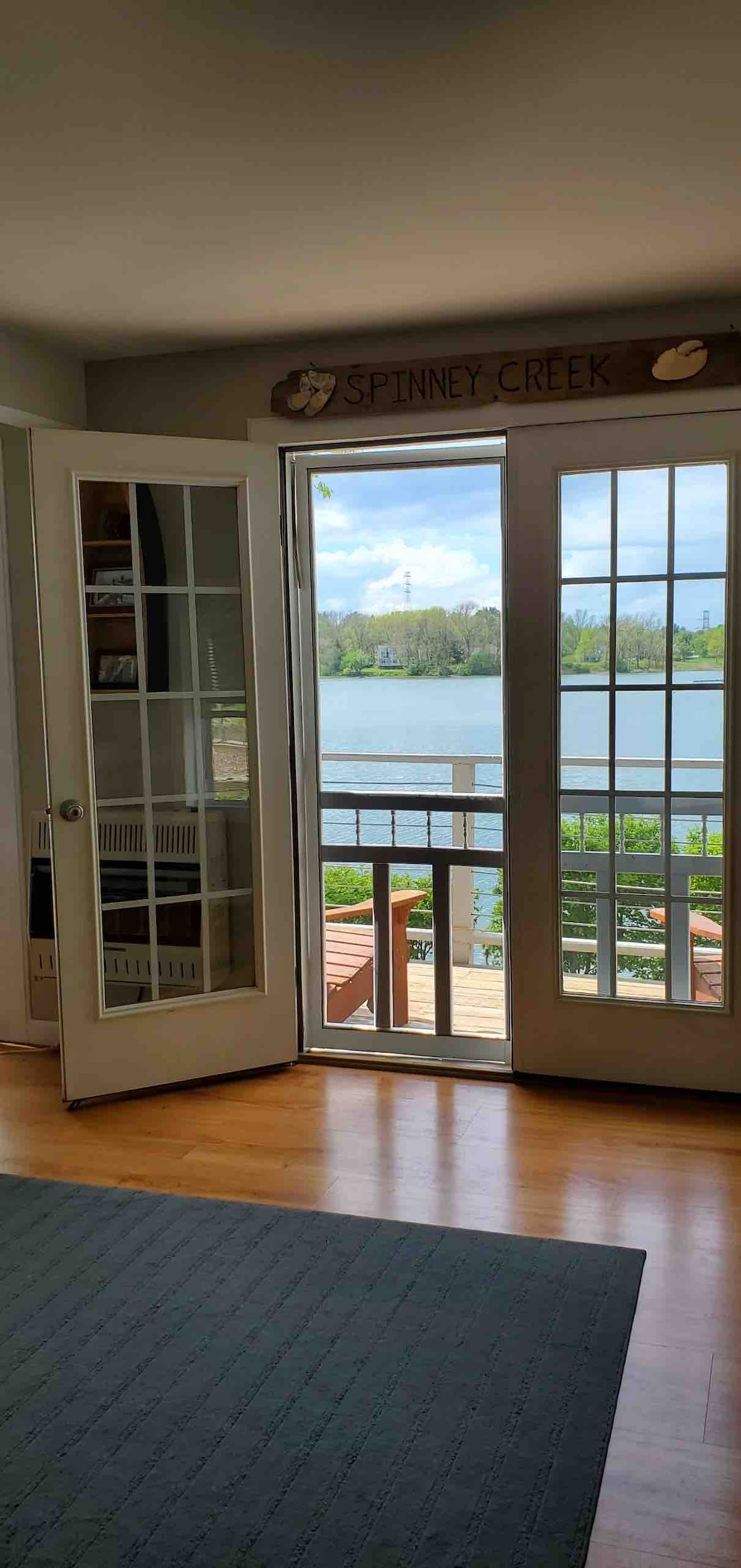 Double French doors are seen open, leading to a waterfront deck. Sunlight filters through glass panels, illuminating the room. A view of Spinney Creek is visible, featuring lush greenery along the shoreline. Light wooden flooring and a blue area rug create a serene interior space.