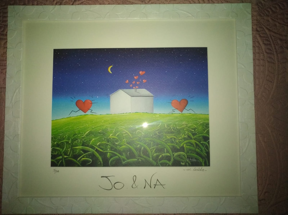 A framed artwork depicts a white house set against a night sky, where a crescent moon and floating hearts are present. The green field in the foreground is contrasted by playful heart figures running towards the house, creating a cheerful and whimsical scene.