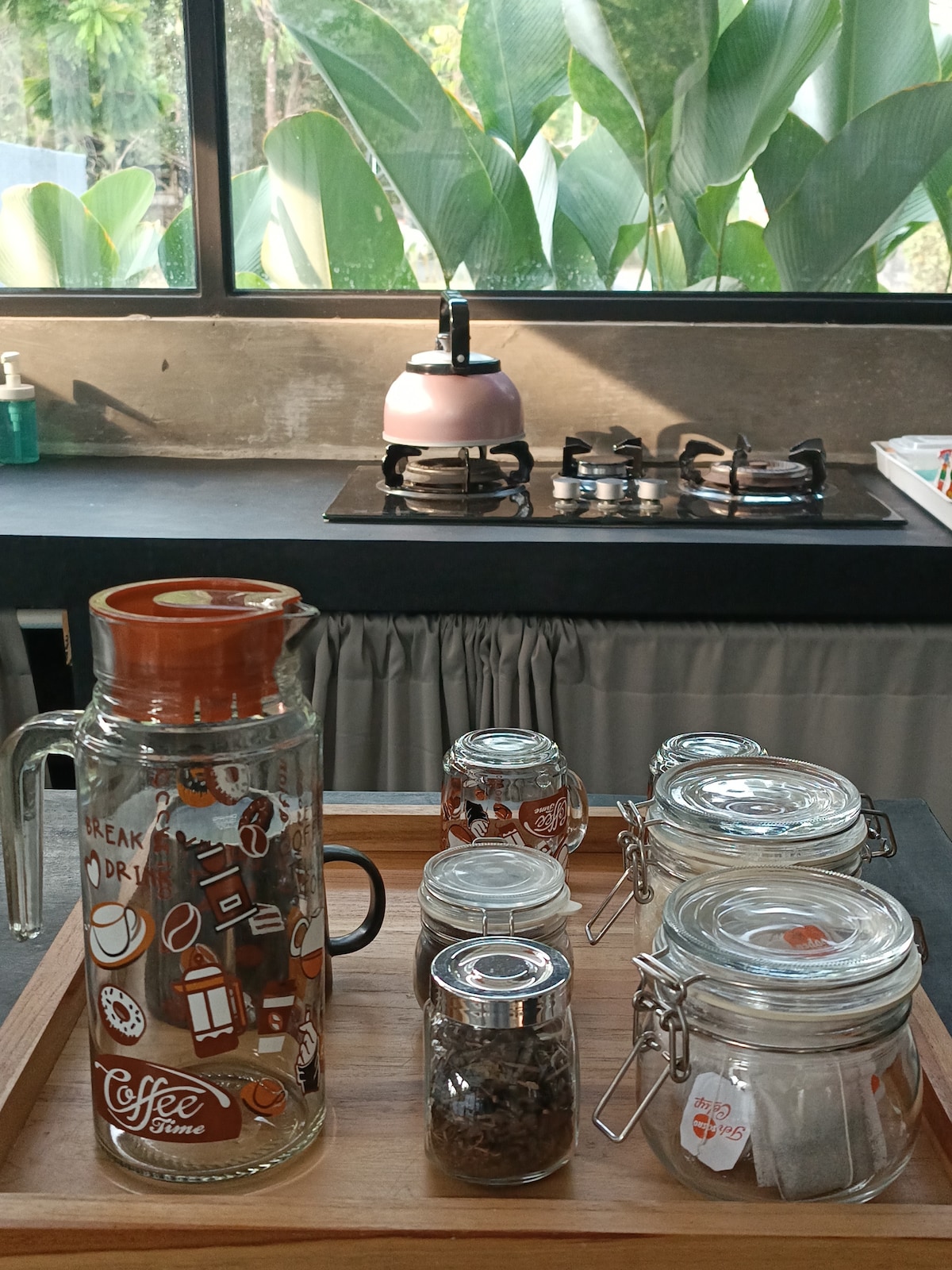 A kitchen area is depicted, featuring a wooden tray with various glass jars and a decorative coffee dispenser. A kettle sits on the stove in the background, with lush greenery visible through large windows, allowing natural light to illuminate the space.