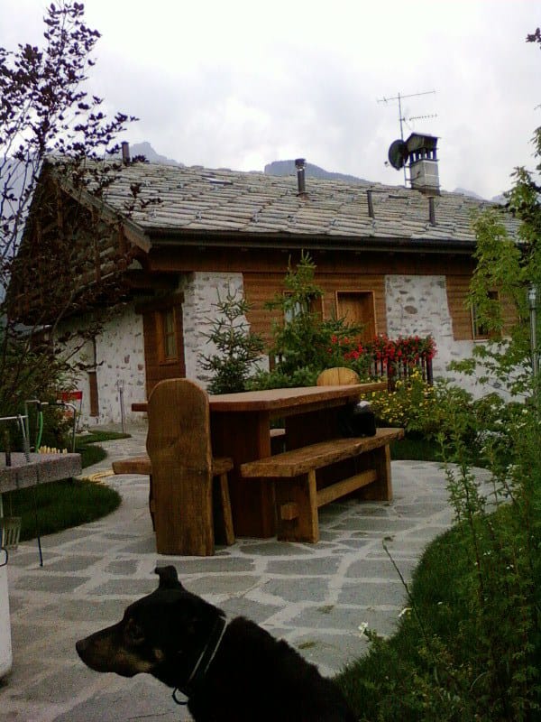 A rustic stone house is framed by lush greenery and blooming flowers. A large wooden table is situated on a stone patio in the foreground, surrounded by nature. A dog is seen resting nearby, adding a homely touch to the peaceful outdoor setting.