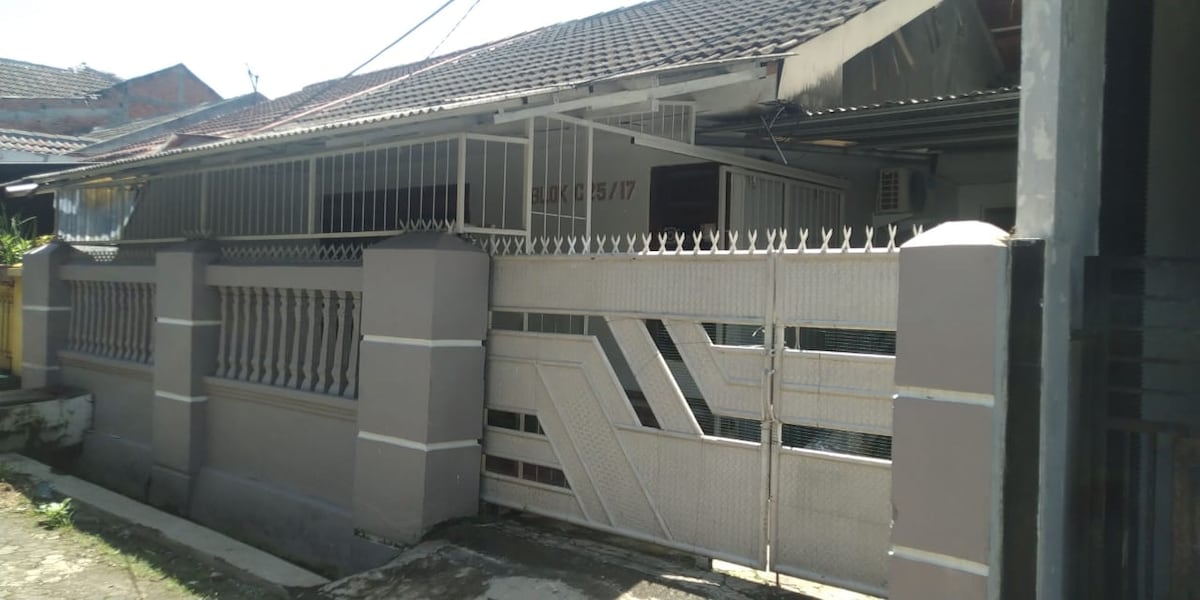 A gated entrance is visible, featuring a sleek metal gate and a solid perimeter wall. The structure showcases a modest single-story design with an overhanging roof, offering protection from the elements. The pathway is paved, leading to the main door of the house.
