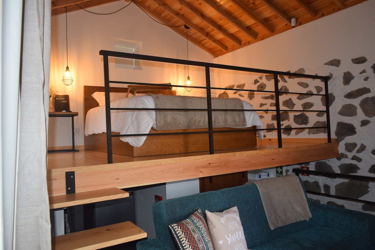 A lofted sleeping area features a spacious bed with neutral-toned linens, illuminated by warm pendant lights. The rustic stone wall adds character, while a cozy seating area below is furnished with a comfortable sofa, inviting relaxation.