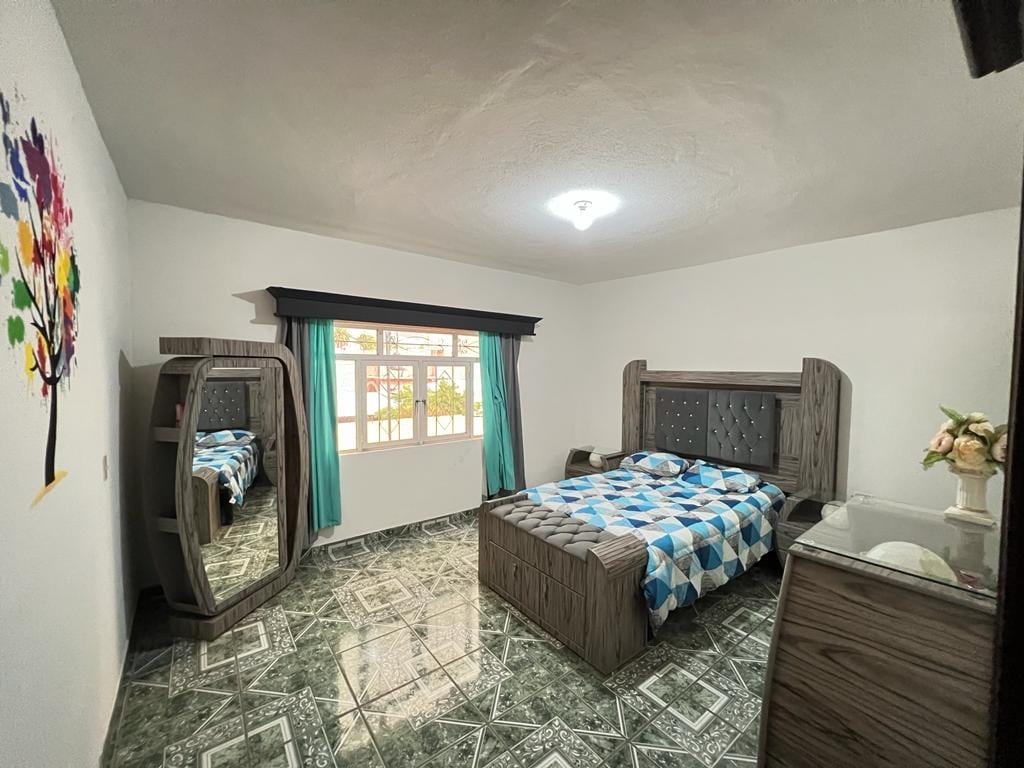 A spacious bedroom is presented with a large bed featuring a patterned blue and white duvet cover. A wooden mirror and a dresser are visible, accompanied by a window adorned with turquoise curtains that allow natural light to fill the room. A colorful mural of a tree adds a decorative touch.