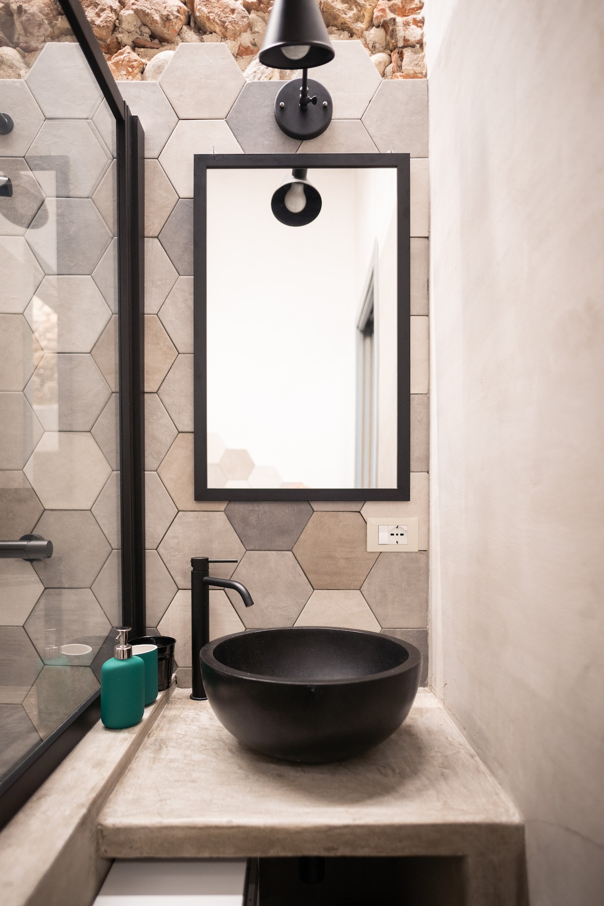 A modern bathroom features a unique round black basin atop a concrete countertop. A rectangular mirror with a black frame hangs above. Textured hexagon tiles create a stylish backdrop, complemented by a minimal wall sconce. A green bottle is positioned nearby.