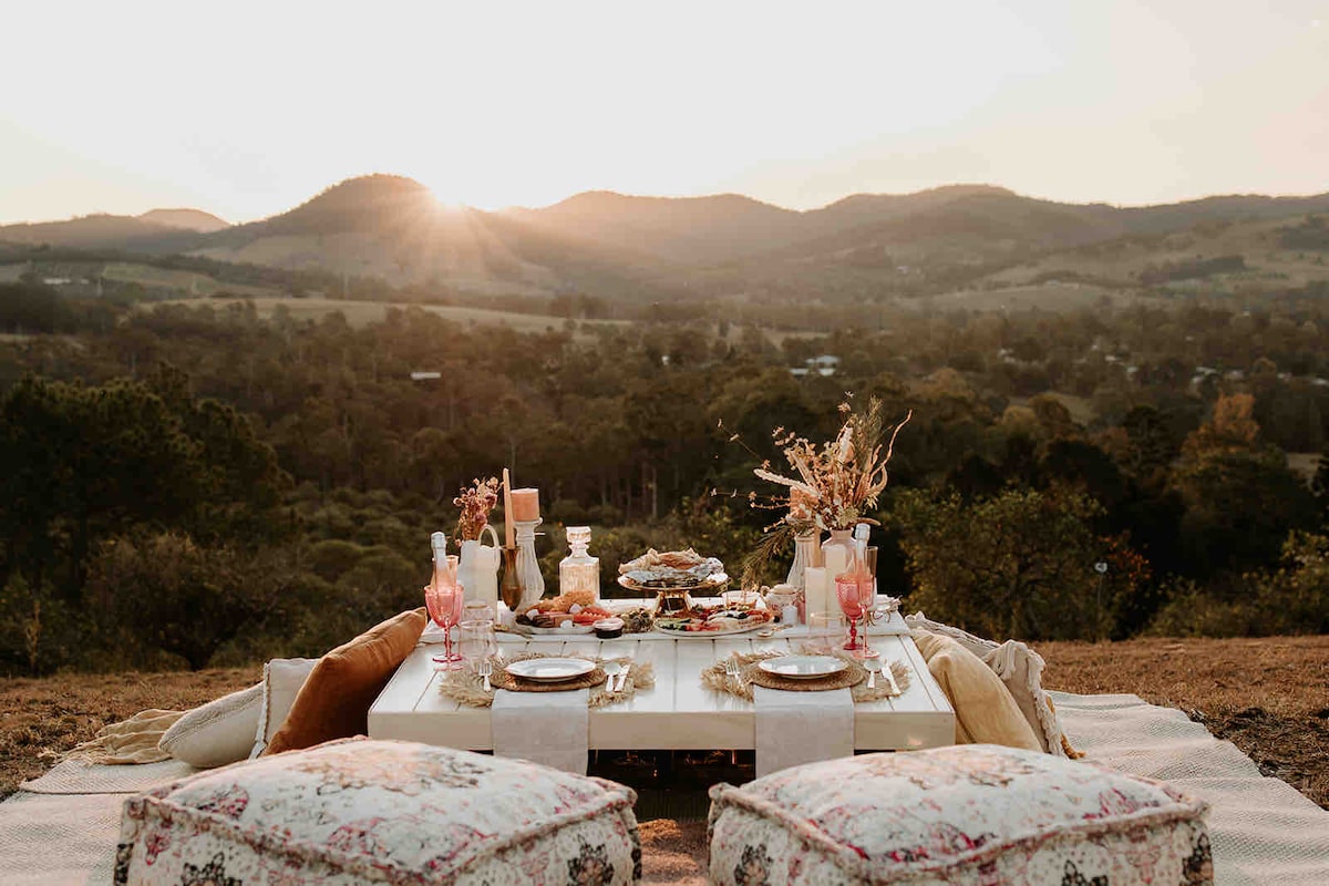 A picnic setup is displayed on a table overlooking rolling hills, with soft cushions providing seating. Plates of delicacies are arranged alongside pink drinks in elegant glasses. The sunset casts a warm glow over the scene, enhancing the natural beauty of the surroundings.