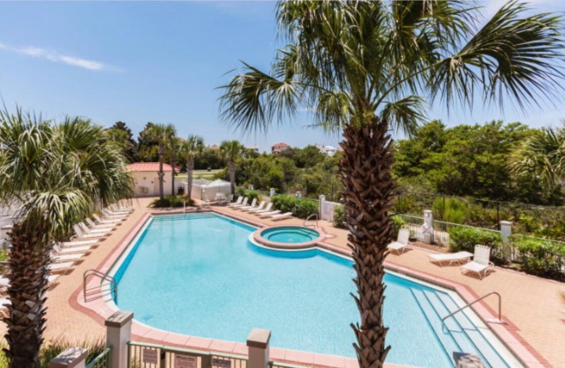 A spacious swimming pool is framed by palm trees, with a hot tub adjacent to it. Numerous lounge chairs are arranged around the pool area on a sunlit deck, providing a serene setting surrounded by greenery.