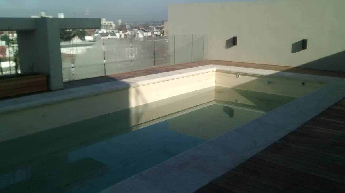 A rooftop pool is presented, featuring clear water surrounded by a wooden deck. Glass partitions provide an unobstructed view of the surroundings, with sunlight creating reflections on the water's surface. The area appears spacious and well-designed for relaxation.