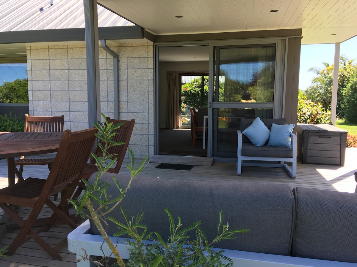 An outdoor seating area is visible, featuring a wooden dining table accompanied by several chairs. A comfortable couch with blue cushions is positioned nearby, enhancing the space's relaxation options. Lush greenery frames the scene, creating an inviting atmosphere for outdoor enjoyment.