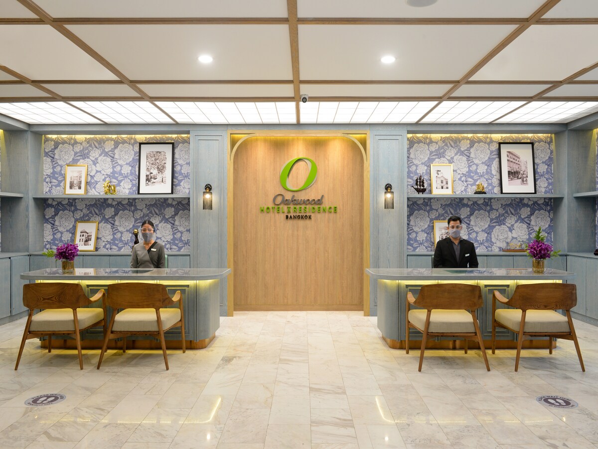The hotel lobby features a welcoming design with a modern reception area. Stylish wooden counters are complemented by decorative blue wallpaper and framed artwork. Potted flowers add a touch of color, enhancing the inviting space for guests upon arrival.