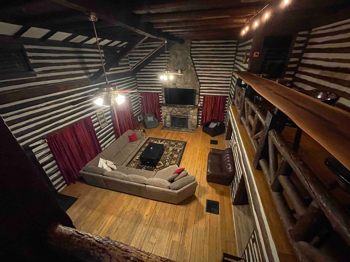 A spacious living area features a large sectional sofa, complemented by accent pillows, and a central coffee table. A stone fireplace is positioned below a mounted TV. Dark wood beams and striped walls create a rustic ambiance, with warm lighting adding to the inviting feel.