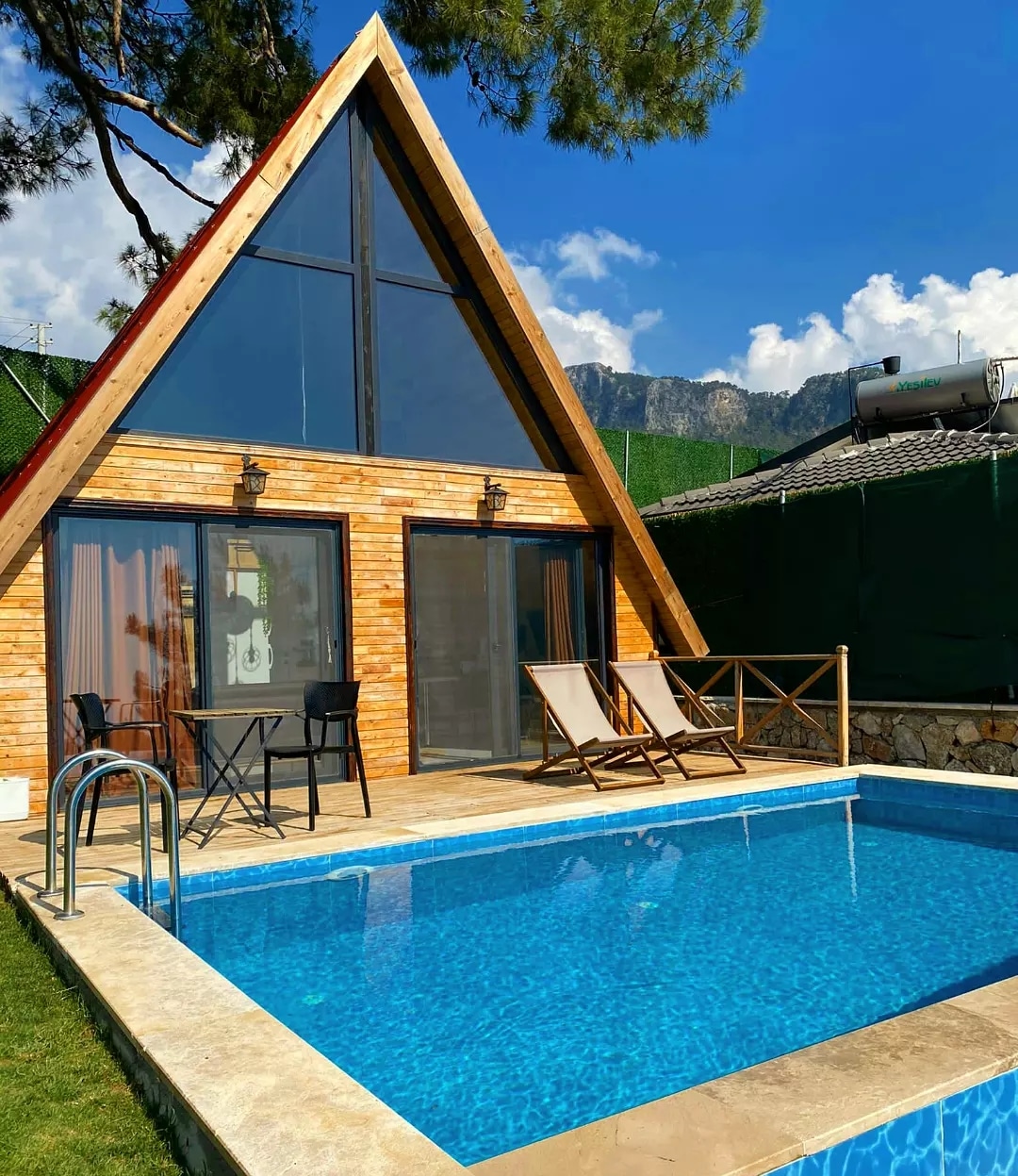 A wooden bungalow with a distinctive triangular roof is situated beside a glistening infinity pool. Two loungers are placed on the deck area, accompanied by a small table. Large glass doors lead into the living space, framed by lush greenery and mountain views.