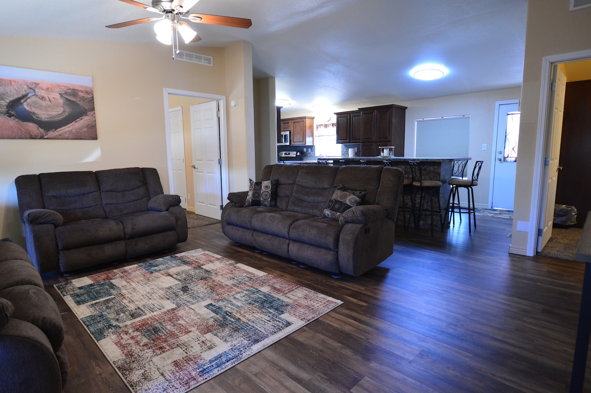 A spacious living area is furnished with two comfortable sofas and a colorful area rug. A ceiling fan provides airflow, while a light fixture illuminates the room. The kitchen and dining area are visible in the background, featuring modern appliances and seating.