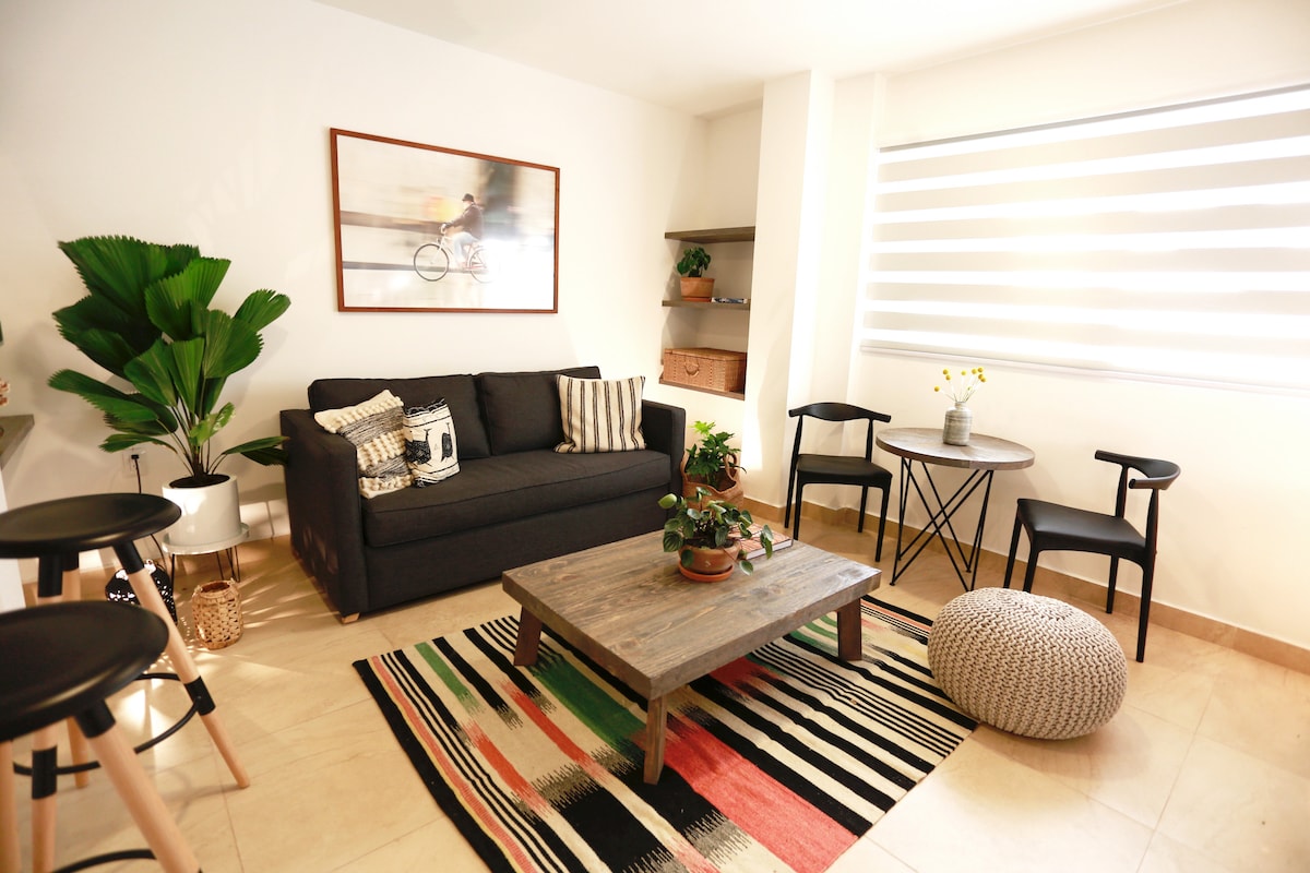 A modern living space is displayed, featuring a dark gray sofa adorned with decorative cushions. A wooden coffee table is centered on a colorful, striped rug. Two chairs and a small table are positioned nearby, with a decorative plant and a vase of flowers adding a touch of greenery.