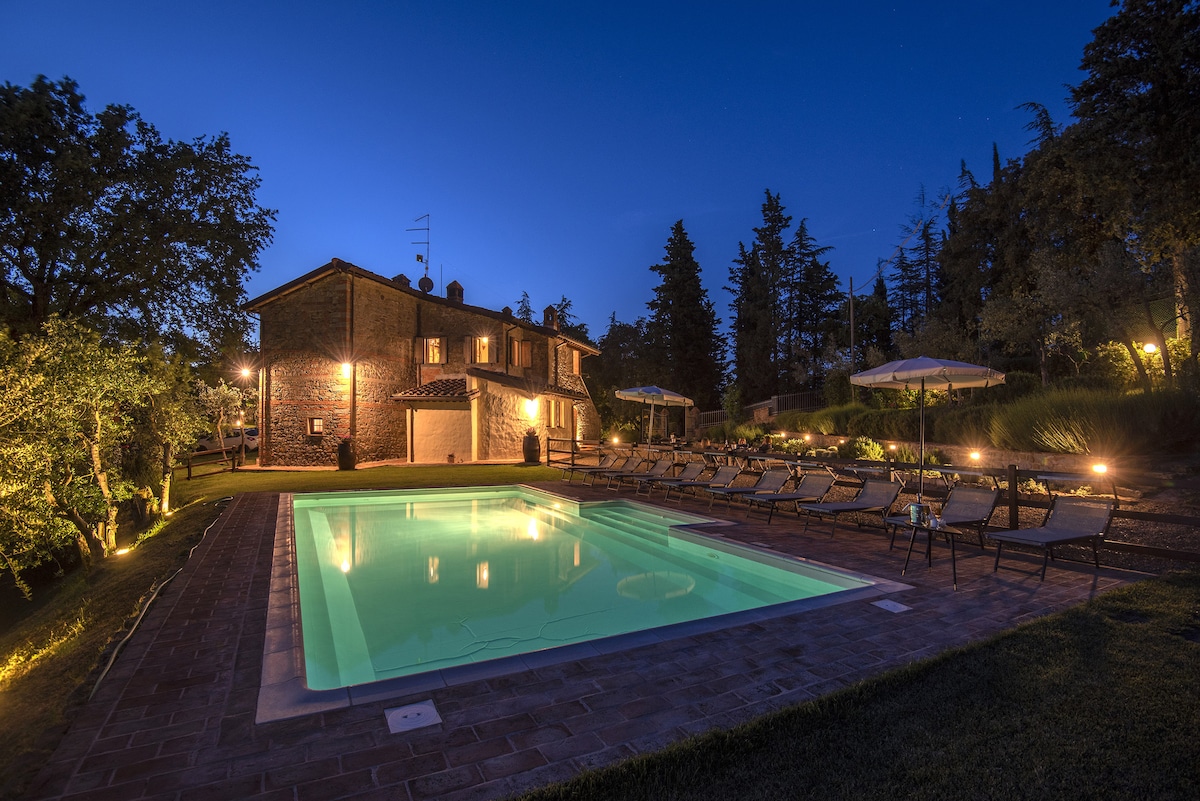 An inviting outdoor space is presented at twilight, featuring a tranquil swimming pool surrounded by lounge chairs and shaded by umbrellas. A rustic stone building serves as a backdrop, illuminated by soft lighting that enhances the evening ambiance. Lush greenery is visible in the surrounding landscape.