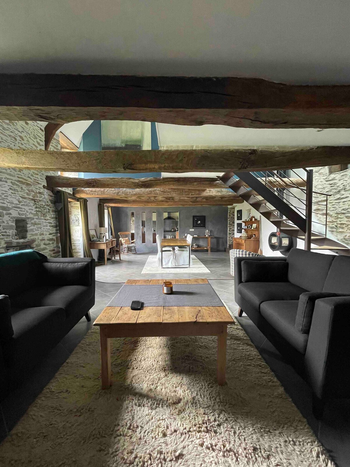 The living area features two black sofas positioned around a wooden coffee table, offering a cozy seating arrangement. Exposed wooden beams and a stone wall add character to the spacious room, which connects to an open layout, illuminated by natural light.