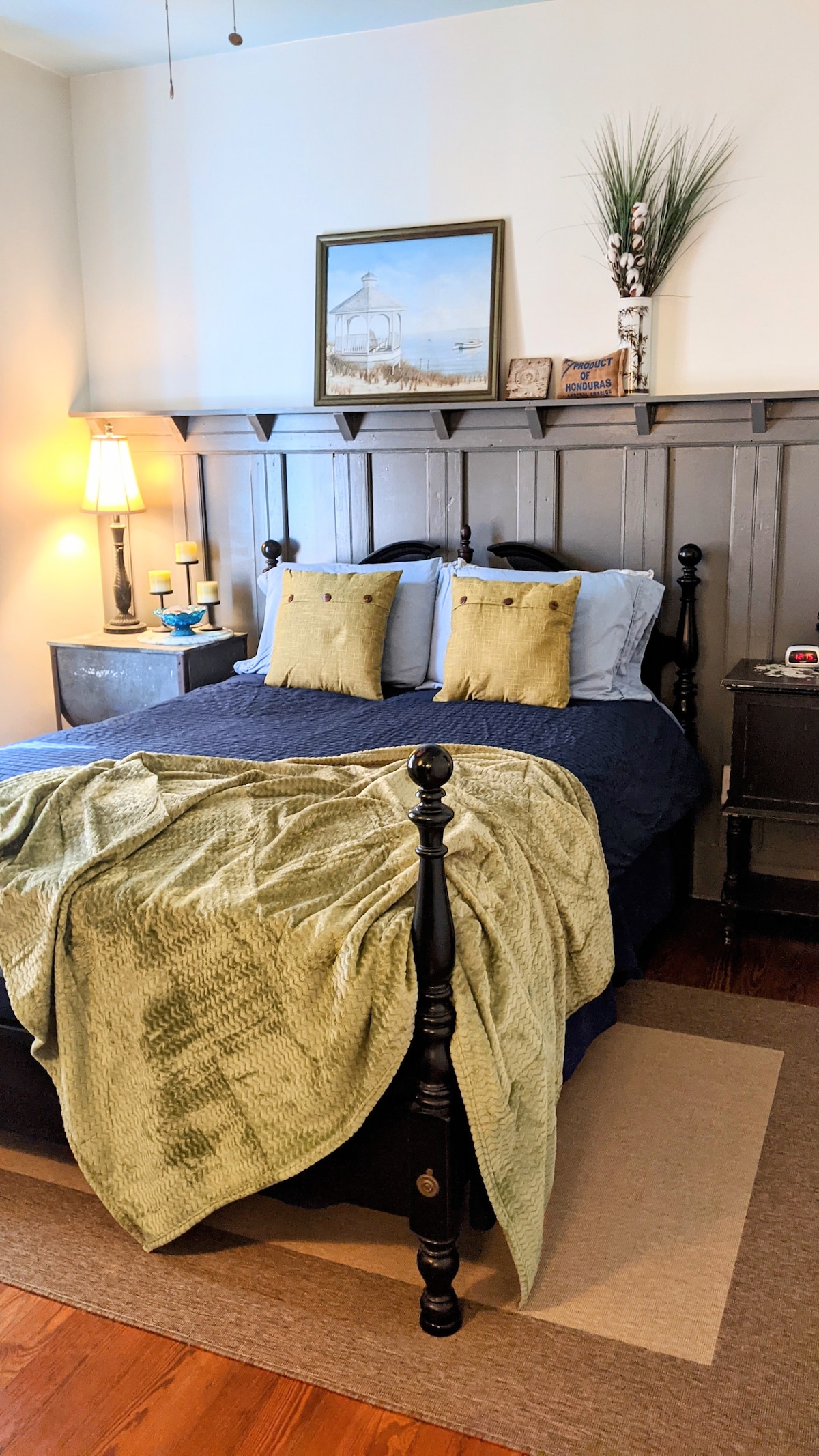A cozy bedroom features a bed with a dark frame and soft linens, accompanied by two decorative pillows. A simple lamp and wall decor add a touch of charm, while a textured green throw is casually draped across the bed.