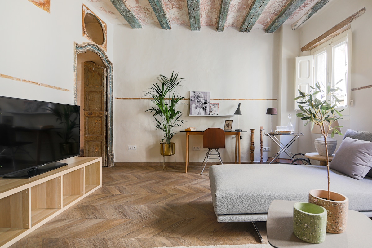 An open living area features a modern sofa positioned next to a flat-screen TV on a wooden console. Natural light fills the space, highlighting the preserved original ceiling beams and decorative doorway. A small workspace with a plant and contemporary chair is visible alongside a chic side table.
