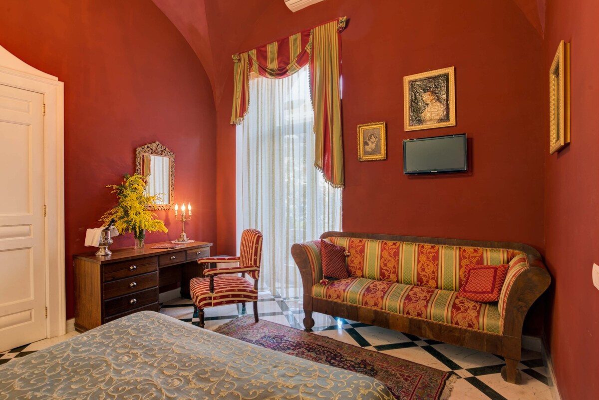 The room features rich red walls and arched ceilings, creating a cozy ambiance. A spacious bed is complemented by a patterned rug. Beside the bed, a wooden dressing table and a chair offer functionality. Natural light filters through sheer drapes, highlighting the exquisite decor.