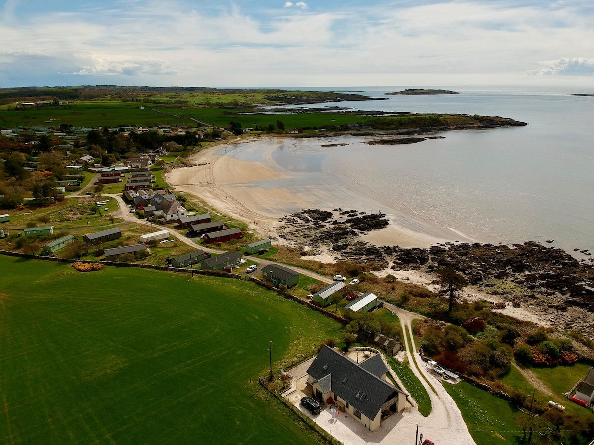 An aerial view captures a coastal landscape featuring a sandy beach and gentle waves lapping against the shore. Green fields and scattered houses line the beach, while distant islands are visible on the horizon. The serene waters reflect the sky's subtle colors.