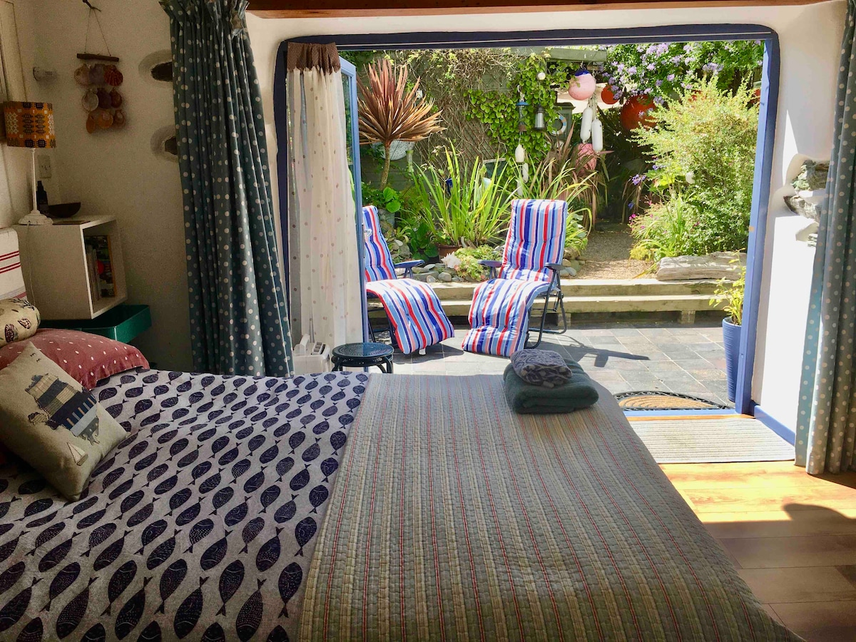 The room features a spacious bed adorned with patterned bedding, positioned near tri-folding doors that open to a private garden space. Two striped lounge chairs are visible outside, accompanied by lush greenery, creating a serene outdoor retreat.