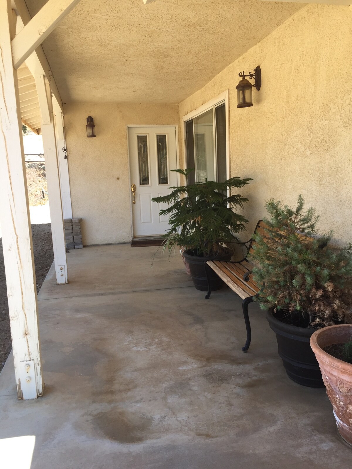 A covered porch area is shown, featuring a wooden bench and potted plants, including a small tree and a shrub. The entrance door is visible, framed by two wall-mounted lanterns, and a smooth concrete surface extends along the porch.