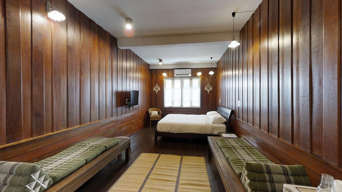 The spacious room features warm wooden paneling and a large window allowing natural light to brighten the space. A queen-sized bed is centrally placed, flanked by two wooden seating areas. A flat-screen TV is mounted on the wall, and a woven rug lies on the wooden floor.