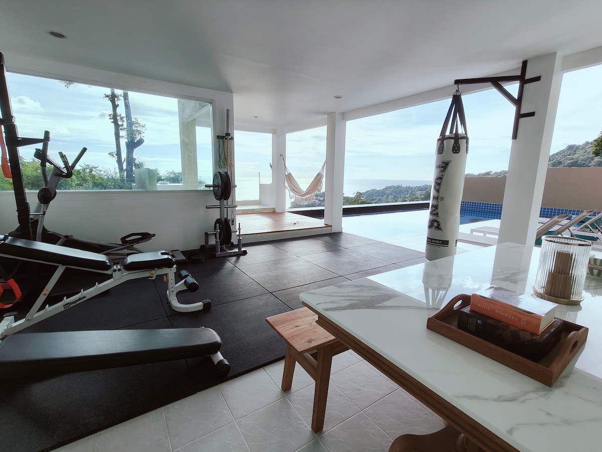 A private fitness area is equipped with exercise machines and a punching bag, offering views of the surrounding landscape. Natural light filters in through large windows, enhancing the open space, while a small table with books adds a touch of comfort.