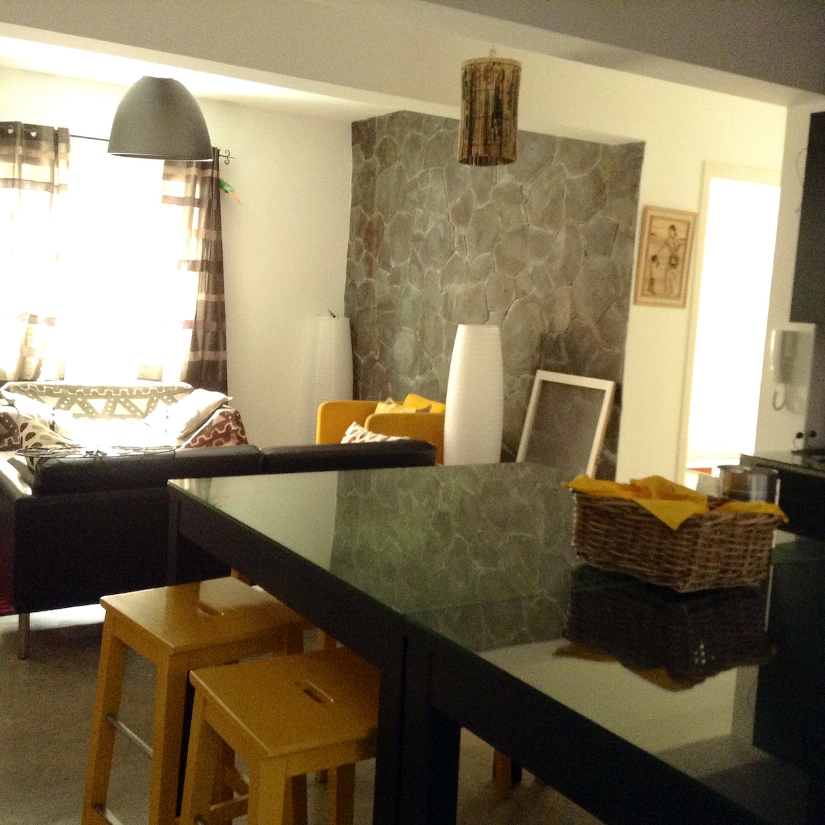 The image depicts a modern living space featuring a large kitchen island with yellow stools. A cozy seating area is visible in the background, adorned with a patterned throw blanket. Natural light enters through the sheer, checkered curtains, enhancing the overall brightness of the room.