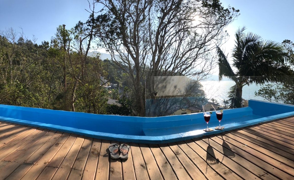 A panoramic view of the coastline is visible from the terrace, framed by lush trees. The wooden floor adds warmth to the space, while two glasses of red beverage are placed on the edge, capturing the light from the sun reflecting off the water.