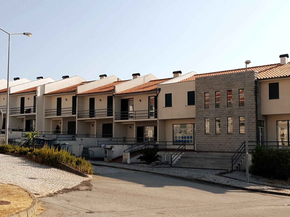 A row of two-story terraced houses features a mix of stone and light-colored façades. Each unit is equipped with a balcony and has black shutters. A paved walkway leads to the main entrance, while plants are visible along the side of the pathway.