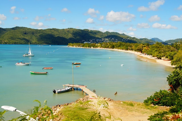 A serene coastal landscape features a calm bay with several colorful boats anchored along the shore. A wooden dock extends into the water, providing a point of access. Lush green hills rise in the background under a partly cloudy sky.