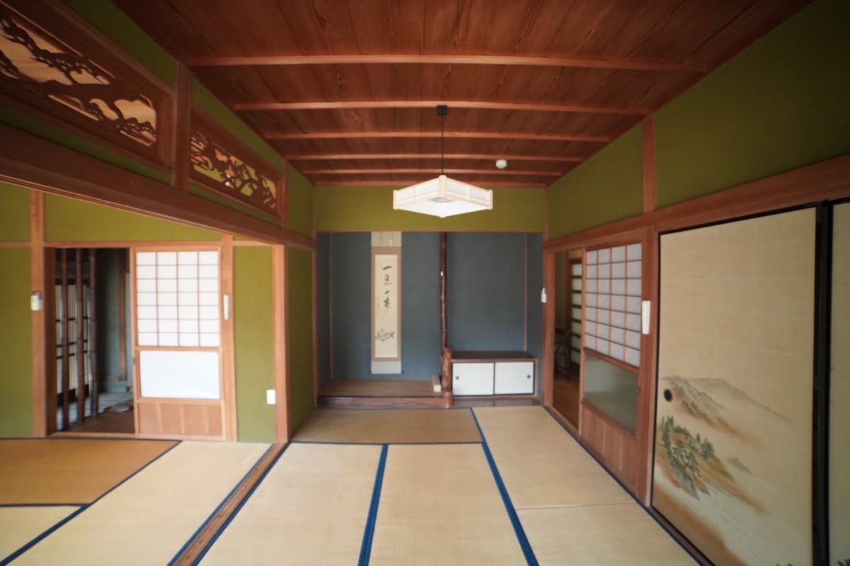 A traditional Japanese room is presented, featuring tatami mat flooring and wooden beams overhead. Sliding doors are visible along the walls, showcasing rice paper panels. The muted green walls are accentuated by woodwork and a central hanging light. A subtle decorative panel enhances the serene atmosphere.