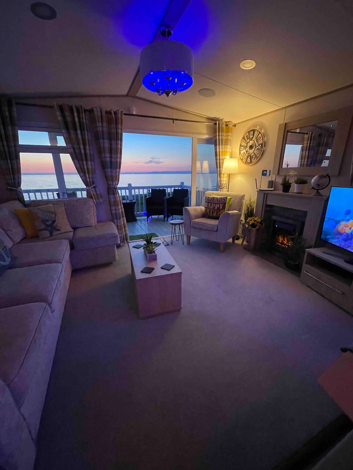 A cozy sitting area is presented with an L-shaped sofa and a single armchair. A coffee table occupies the center, surrounded by soft lighting. Large windows offer a view of the sunset, while a fireplace adds warmth to the space. Decorative elements enhance the atmosphere.