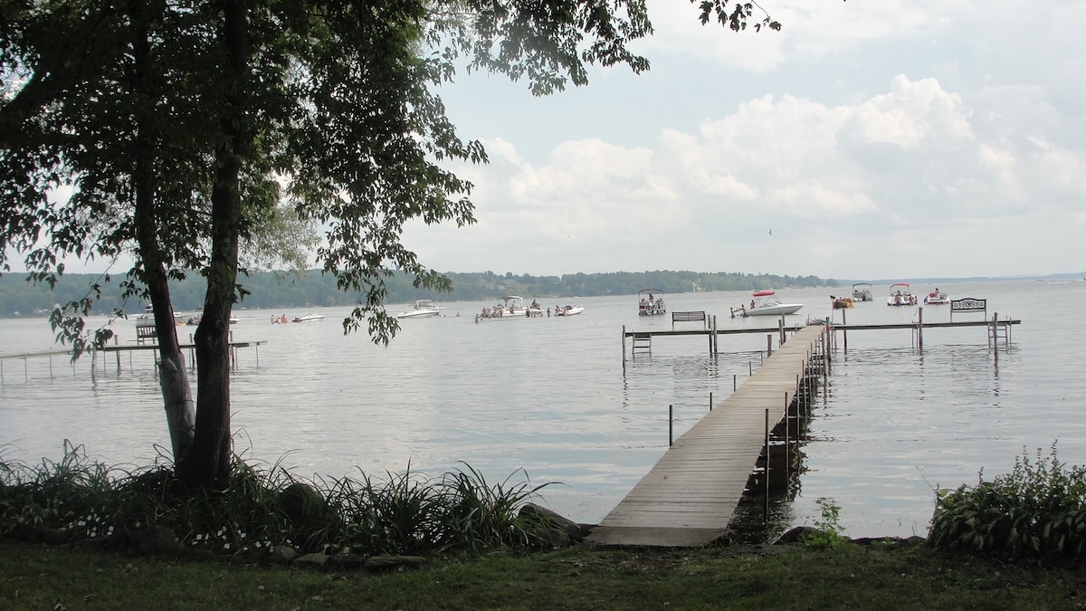 A wooden dock extends into the water, providing access for boats. The peaceful lake is visible, with light ripples reflecting clouds overhead. Green foliage frames the scene, adding to the natural setting. Boaters can be seen enjoying the water in the distance.