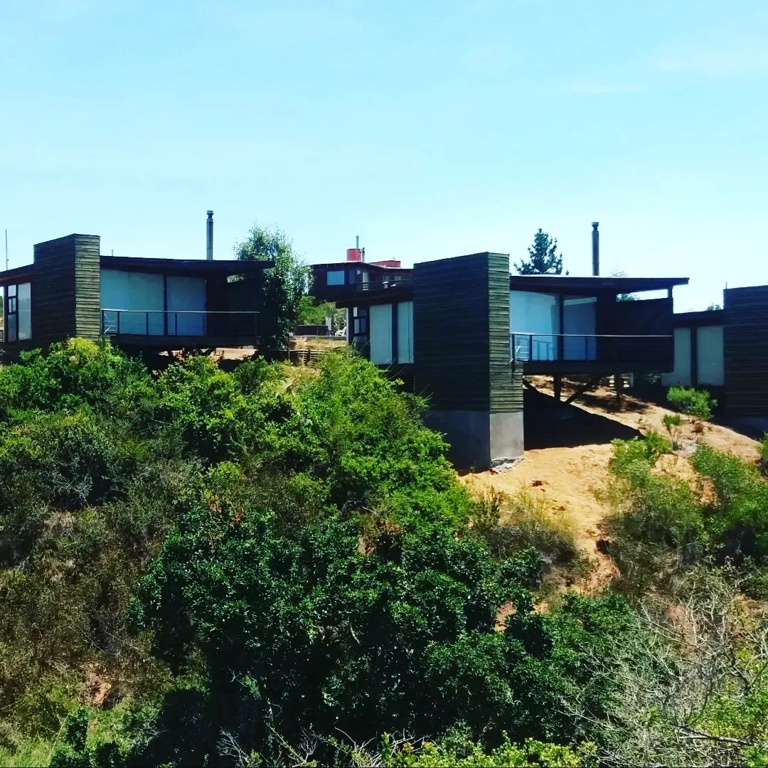 Two modern cabins are positioned on a gently sloping hillside, surrounded by lush greenery. Each cabin features large windows that offer views of the natural landscape. The structure is set apart, providing privacy and a serene environment.