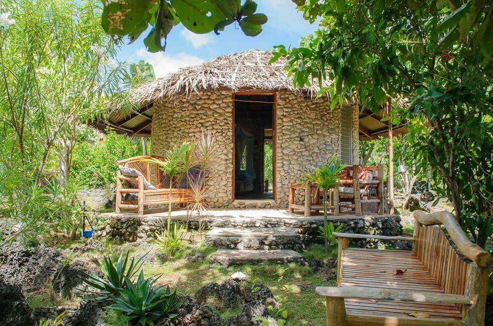 A round, natural stone house is surrounded by lush greenery, with a thatched roof blending harmoniously with the landscape. A wooden seating area is positioned nearby, offering a serene spot to enjoy the garden's tranquility.